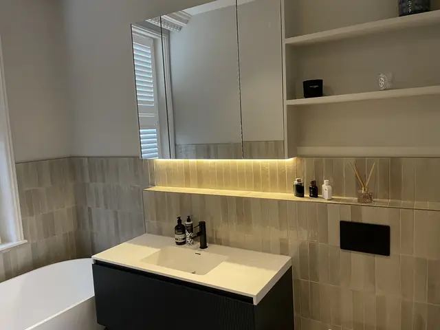 Modern bathroom vanity with dark ribbed cabinet, white sink, illuminated mirror, and built-in shelving.