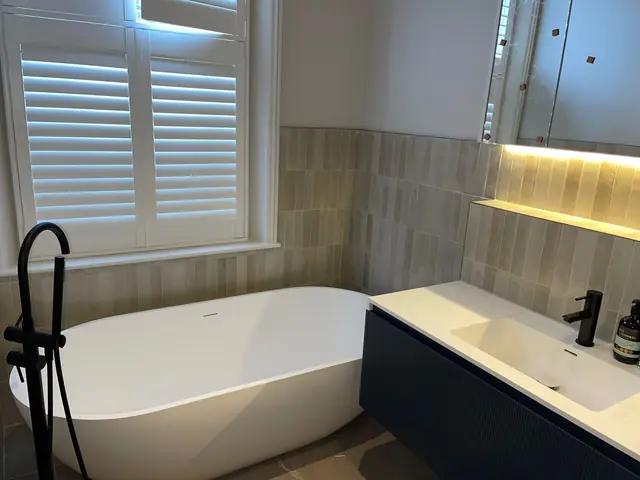 Modern bathroom with freestanding white bathtub, black fixtures, and dark blue vanity with white countertop and illuminated mirror.