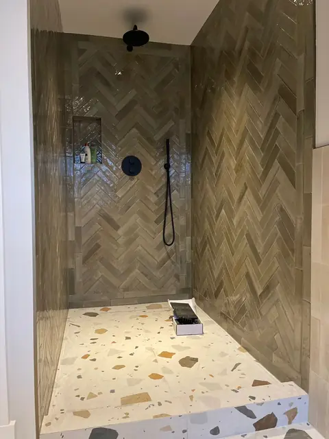 Walk-in shower with elegant herringbone tile pattern in neutral tones and terrazzo flooring, featuring black modern fixtures.