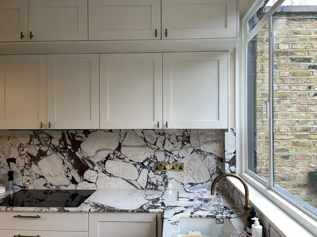Kitchen sink area with white cabinets, luxurious marble splashback, brass tap, and window with brick view.