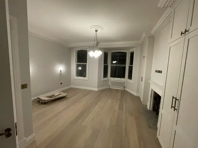 Empty living room with light wood flooring, white fireplace, and elegant bay windows.
