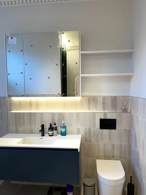 Modern bathroom featuring a white sink on a dark blue vanity, large illuminated mirror cabinet, and white toilet with grey tiled walls.