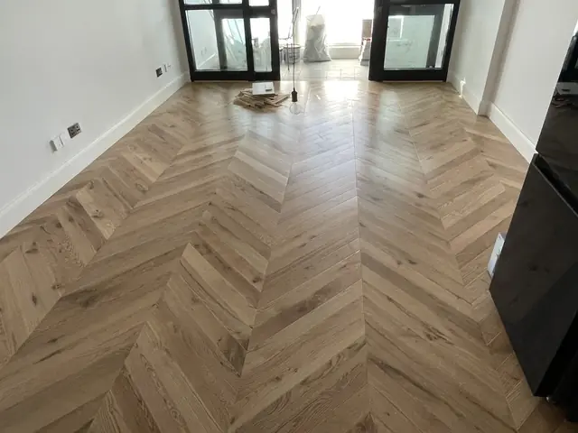 Light oak herringbone parquet wood flooring in a bright, empty space.