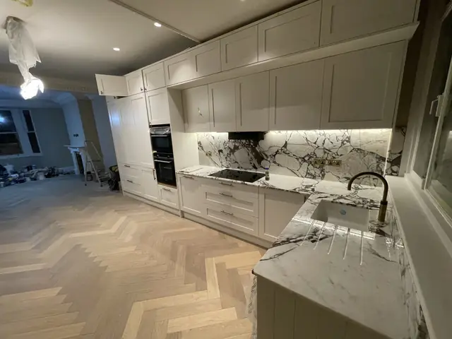 Elegant white kitchen with marble worktops and herringbone flooring during final stages.